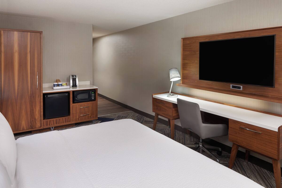 A hotel room with a large bed, a desk with a chair, a television mounted on the wall, a wardrobe, and a kitchenette with a coffee maker and microwave.