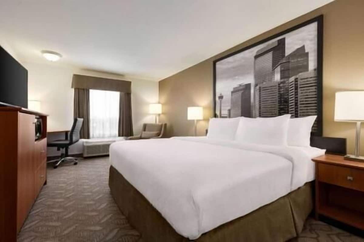 A hotel room with a large bed, a desk, a chair, two lamps, and a cityscape painting on the wall.