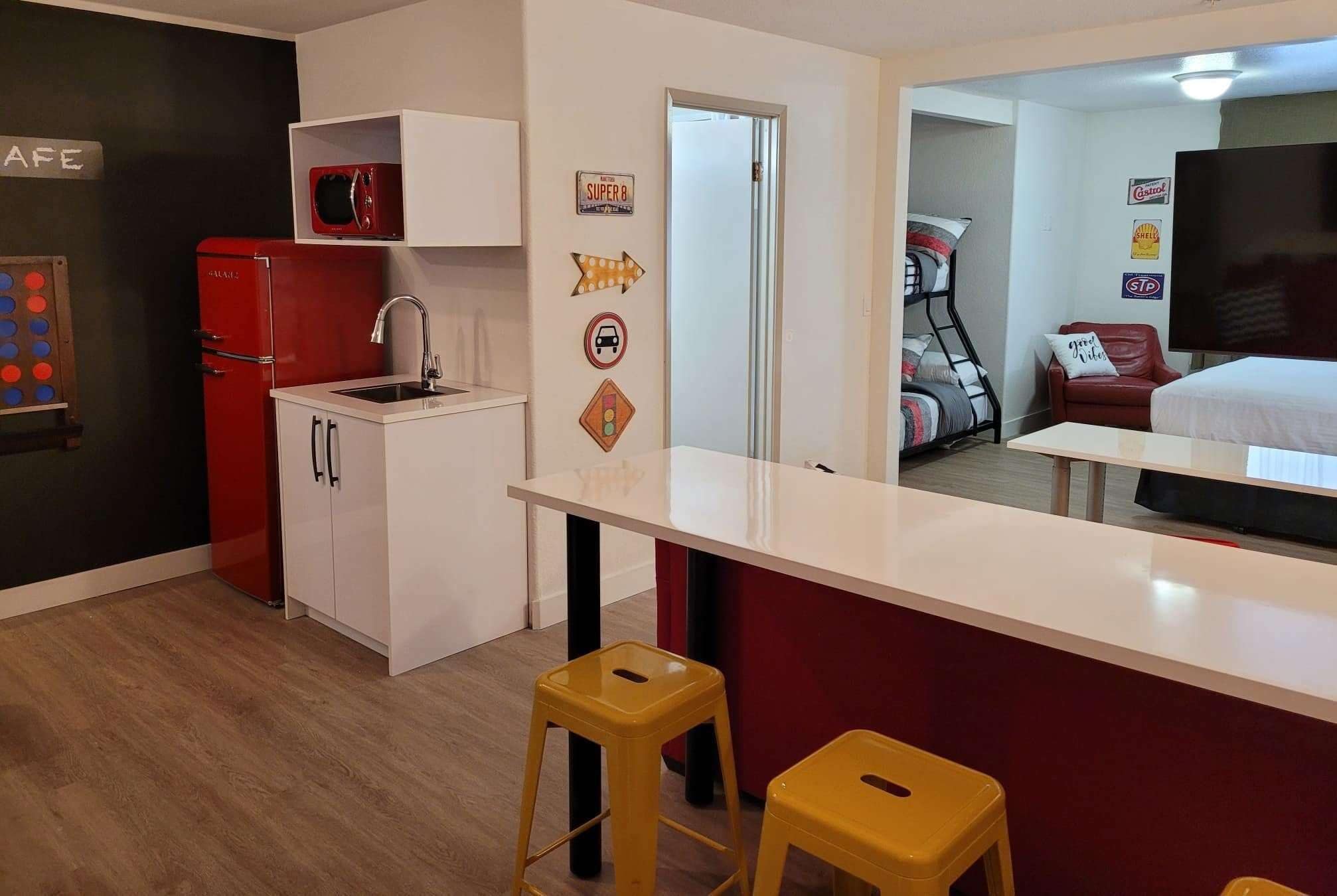 A compact kitchen with a red refrigerator, white cabinets, and a microwave. There are two yellow stools and a large mirror reflecting the room.