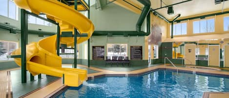 An indoor swimming pool with a yellow water slide and a view of the outside through large windows.