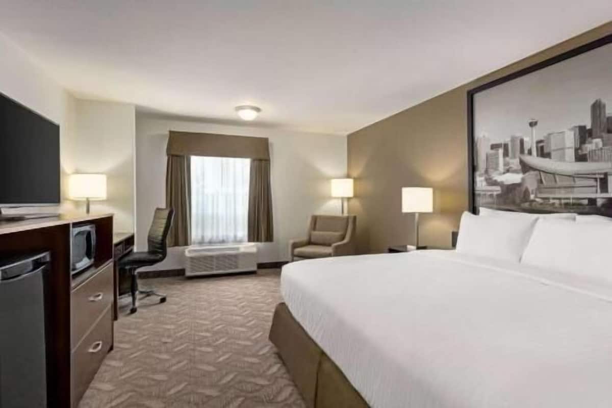 A hotel room with a bed, a desk with a TV, two chairs, a window with curtains, and a cityscape painting on the wall.
