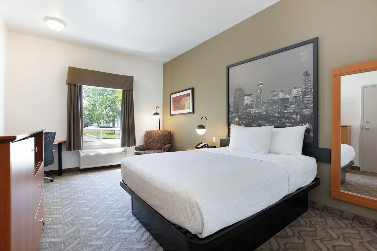 A hotel room with a large bed, a desk, a chair, a cityscape painting, and a window with curtains.