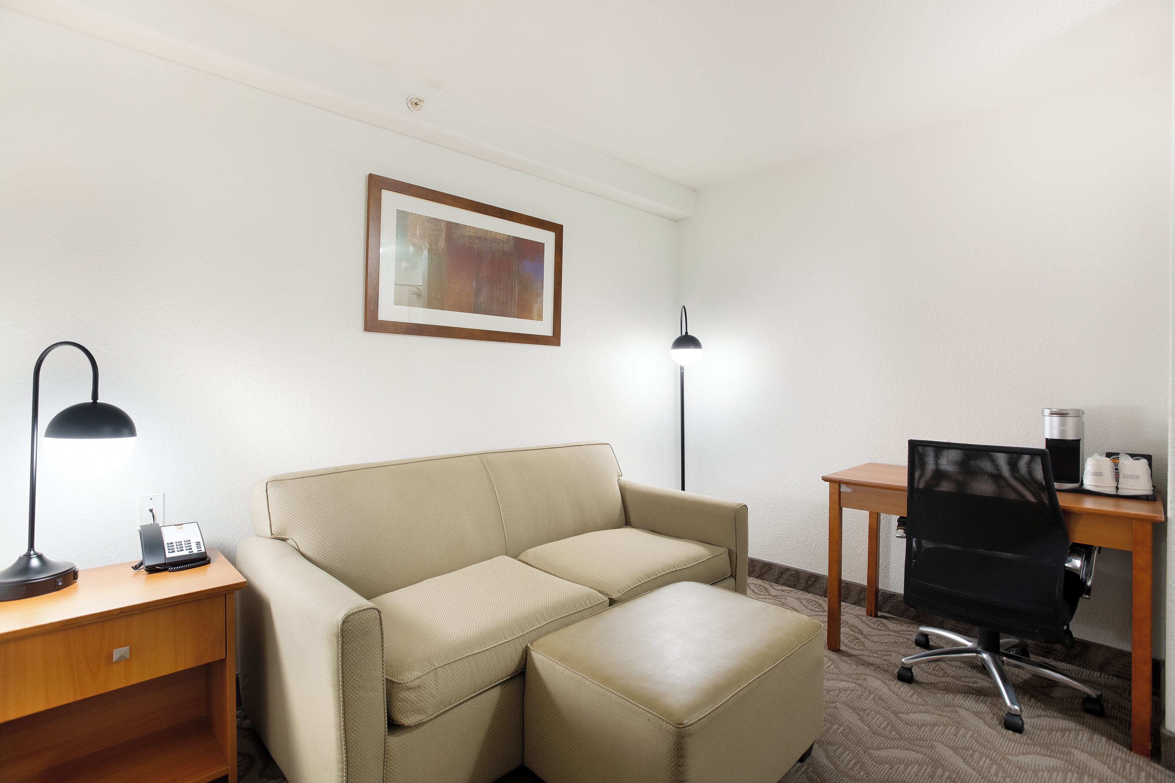 Une chambre d’hôtel avec un canapé beige, un bureau avec une lampe, un ordinateur et un tableau encadré au mur.