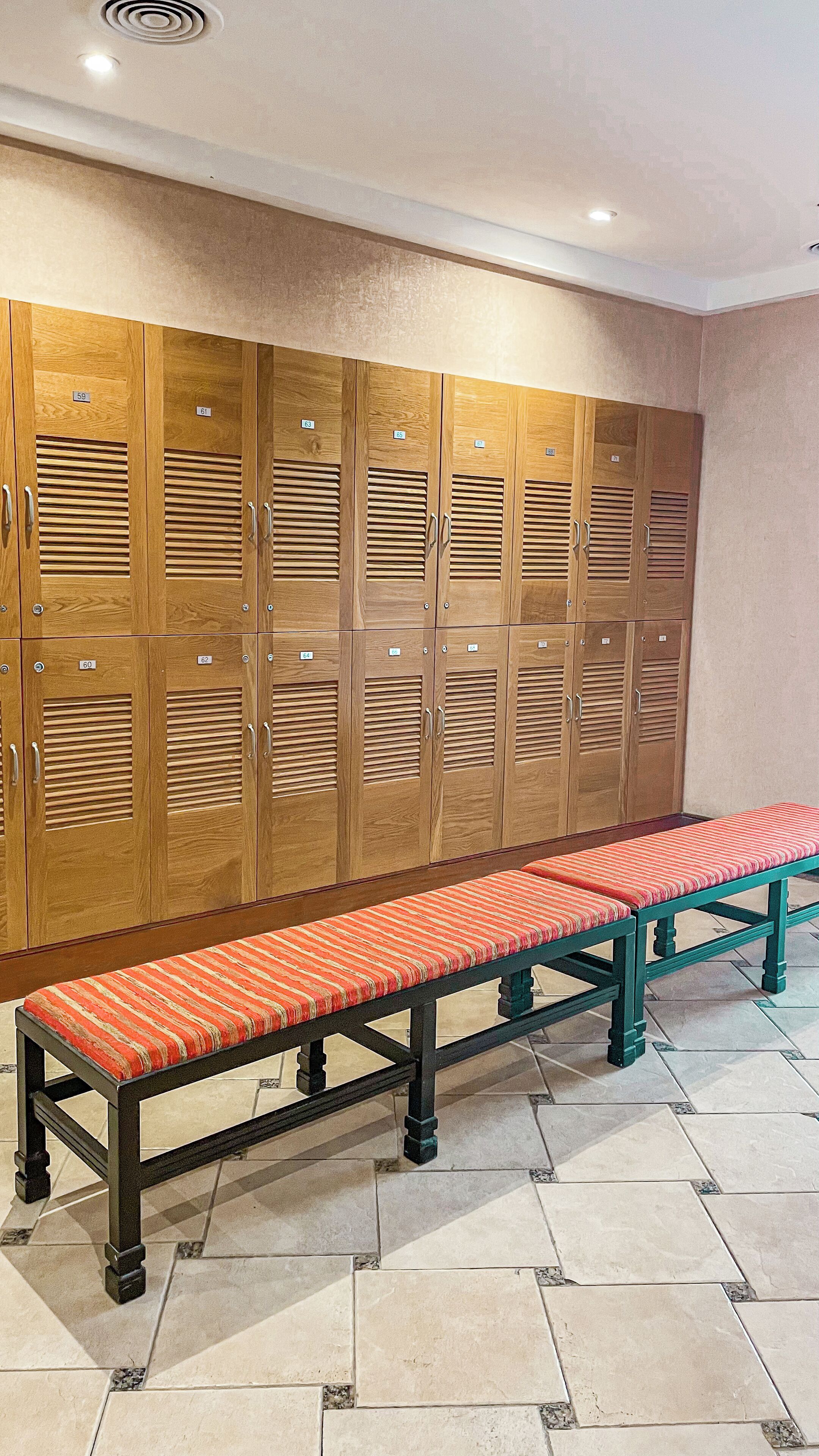 A row of wooden lockers with numbered tags, a bench with a striped cushion, and a tiled floor.