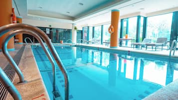 An indoor swimming pool with clear water, metal handrails, and lounge chairs by the poolside.