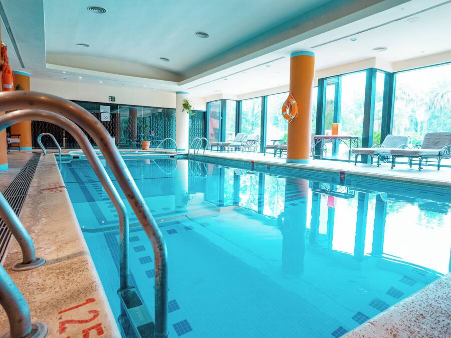 An indoor swimming pool with clear water, metal handrails, and lounge chairs by the poolside.