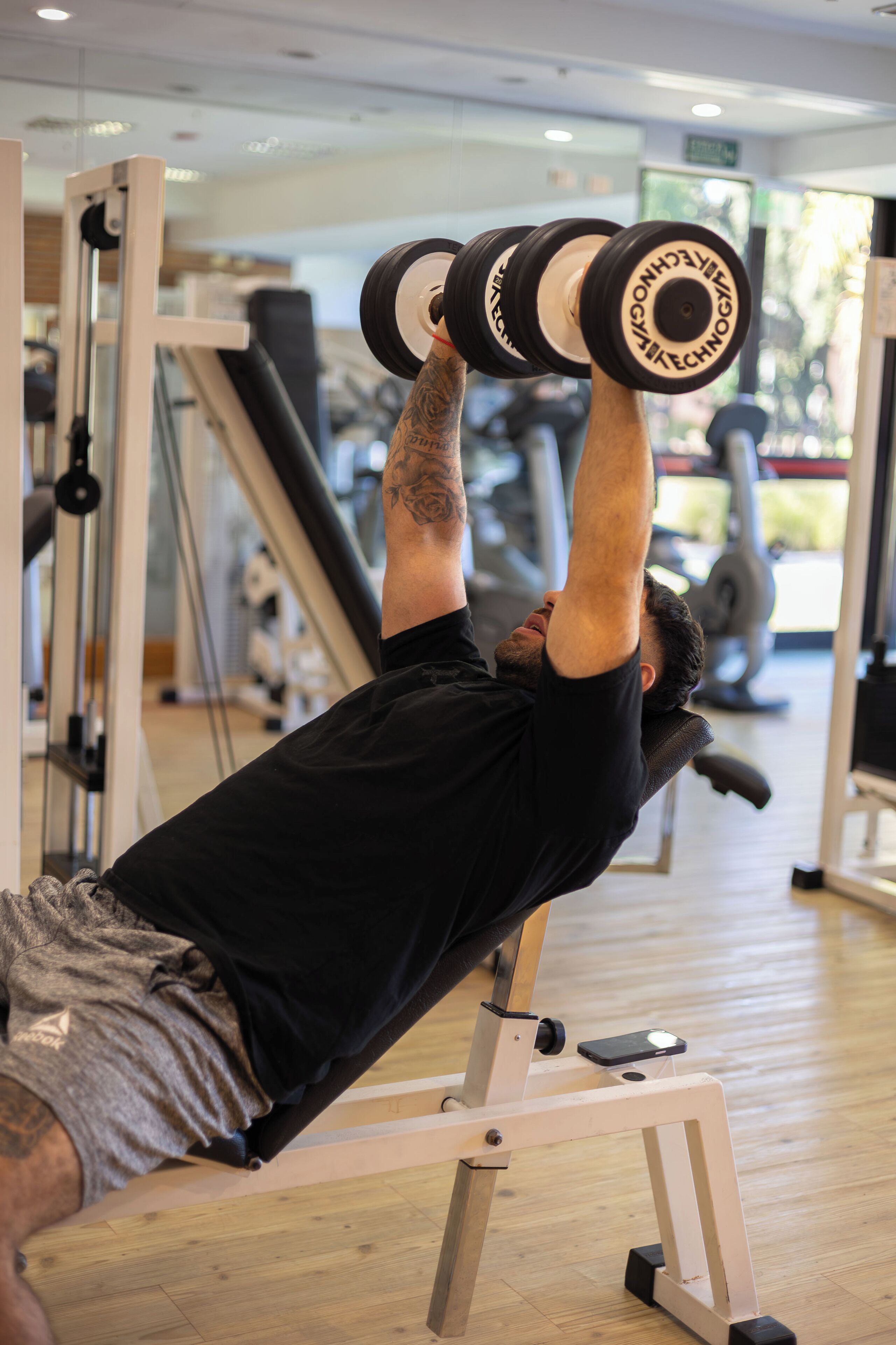 Una persona acostada en un banco dentro de un gimnasio, realizando un ejercicio de hombros con mancuernas.