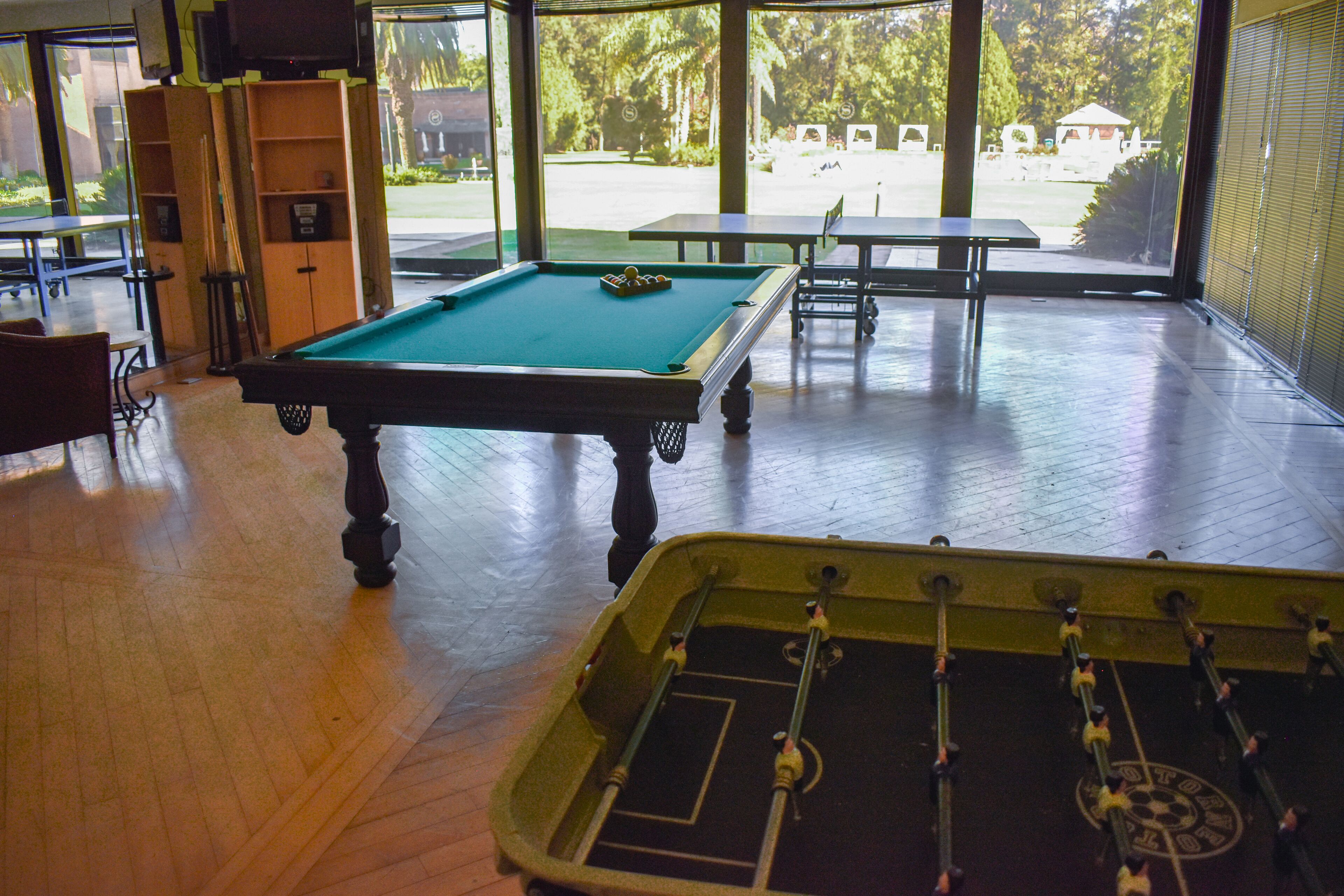 Una mesa de billar en una habitación con una mesa de fútbol de mesa, suelo de madera y ventanas grandes que ofrecen vistas al exterior.