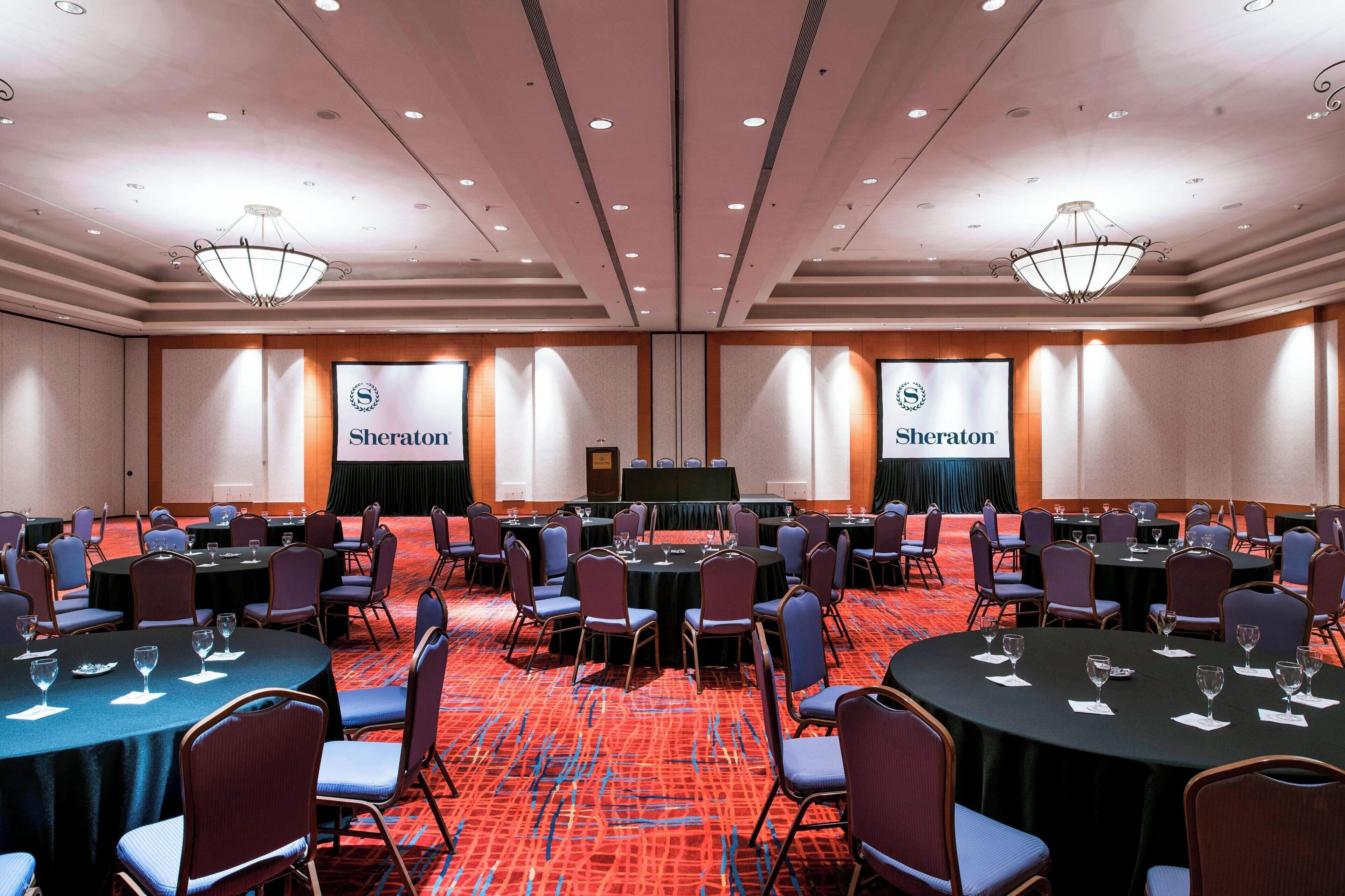 A conference room with multiple tables and chairs, a large screen, and Sheraton branding on the walls.
