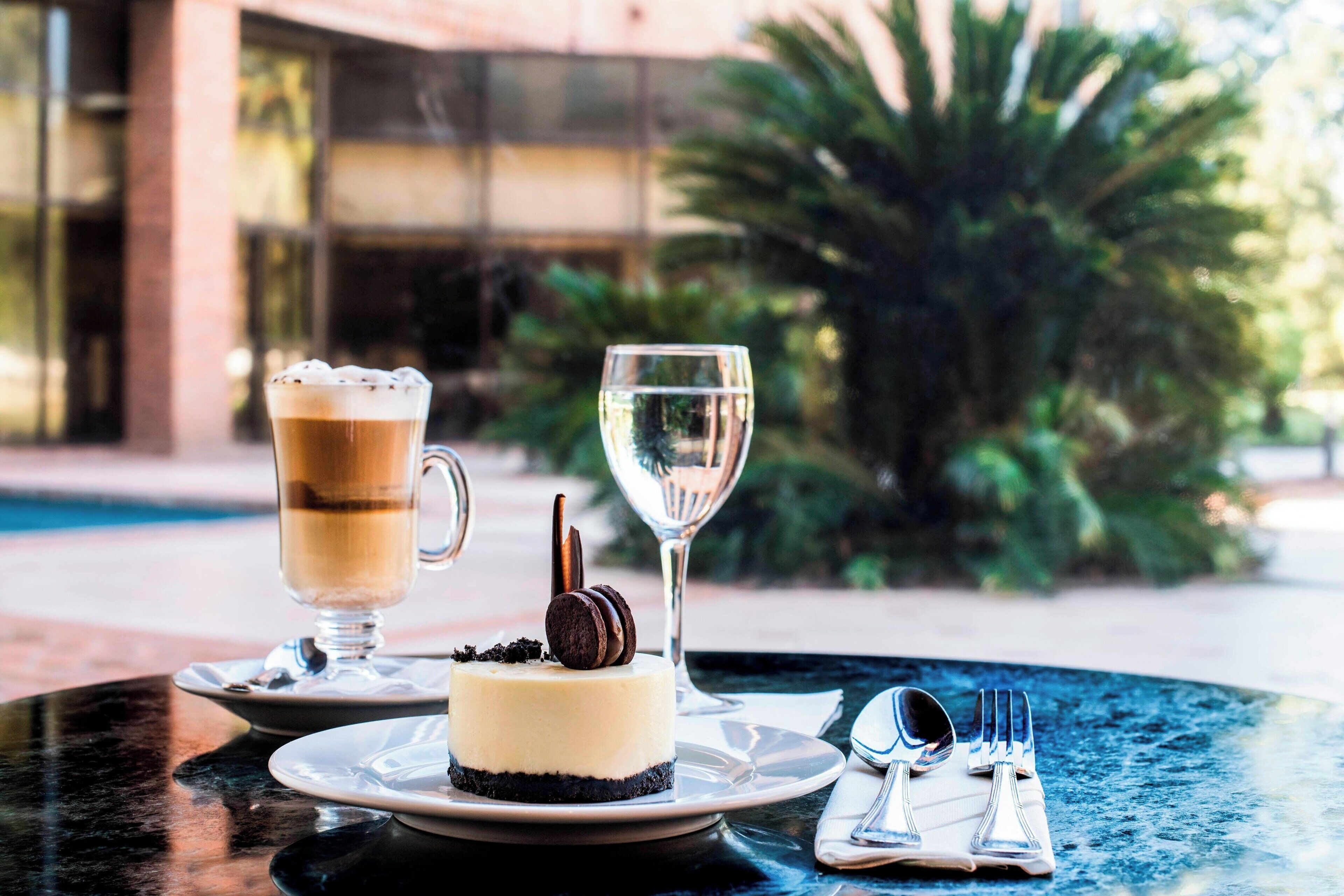 Ein Desserttisch mit Käsekuchen, einer Kaffeetasse und einem Weinglas, aufgestellt an einem Tisch im Freien mit Blick auf ein Gebäude und Grünflächen im Hintergrund.