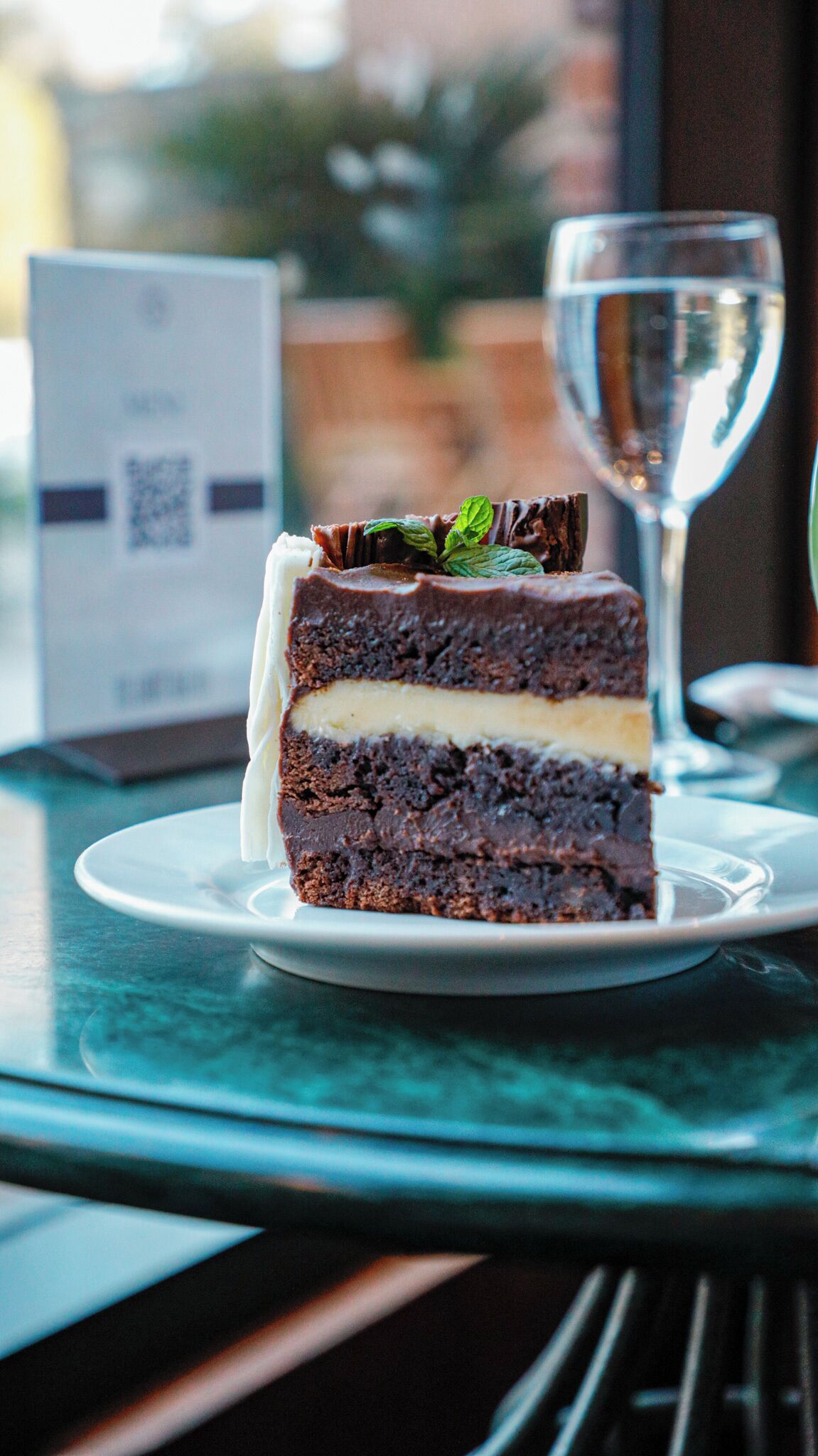 En bit chokladtårta med lager av vit grädde, serverad på en vit tallrik, tillsammans med ett glas vatten, på ett bord med en QR-kod i bakgrunden.