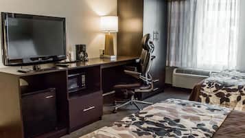 Habitación de hotel con televisión de pantalla plana, escritorio con lámpara, una silla, cama con colcha estampada y ventana con cortinas.