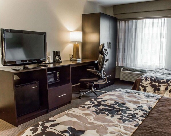 A hotel room with a flat-screen TV, a desk with a lamp, a chair, a bed with a floral bedspread, and a window with curtains.
