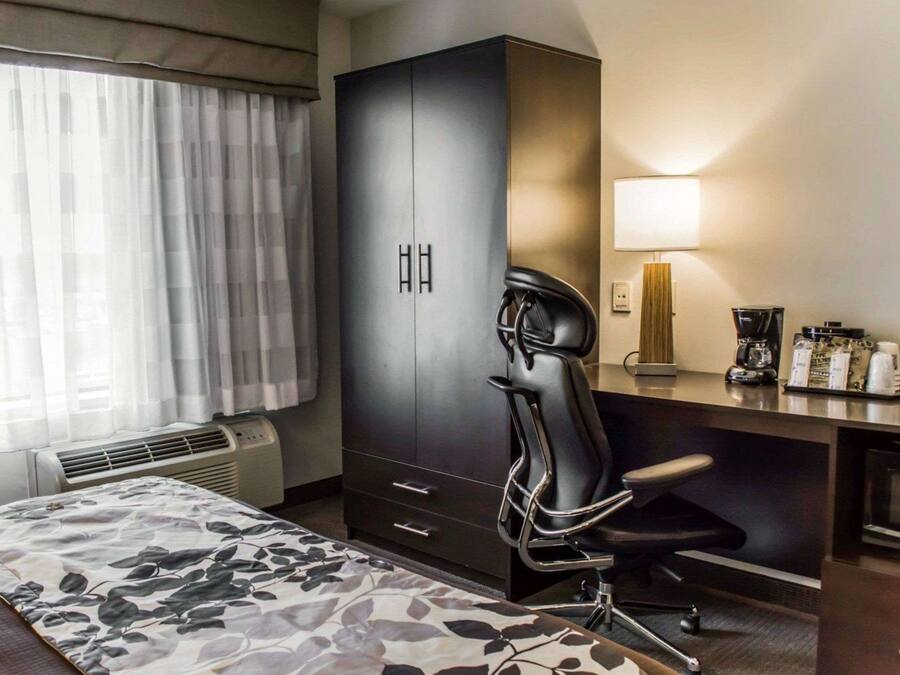 A hotel room with a bed, a desk with a lamp, a chair, a wardrobe, and a coffee machine.