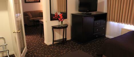 A hotel room with a bed, a television, a dresser, a small table with a vase of flowers, a mirror, and a door leading to another room.