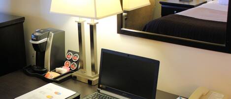 Ein Hotelzimmer mit Schreibtisch, auf dem ein Laptop, eine Kaffeemaschine, ein Telefon und ein Spiegel zu sehen sind, der das Zimmer reflektiert.