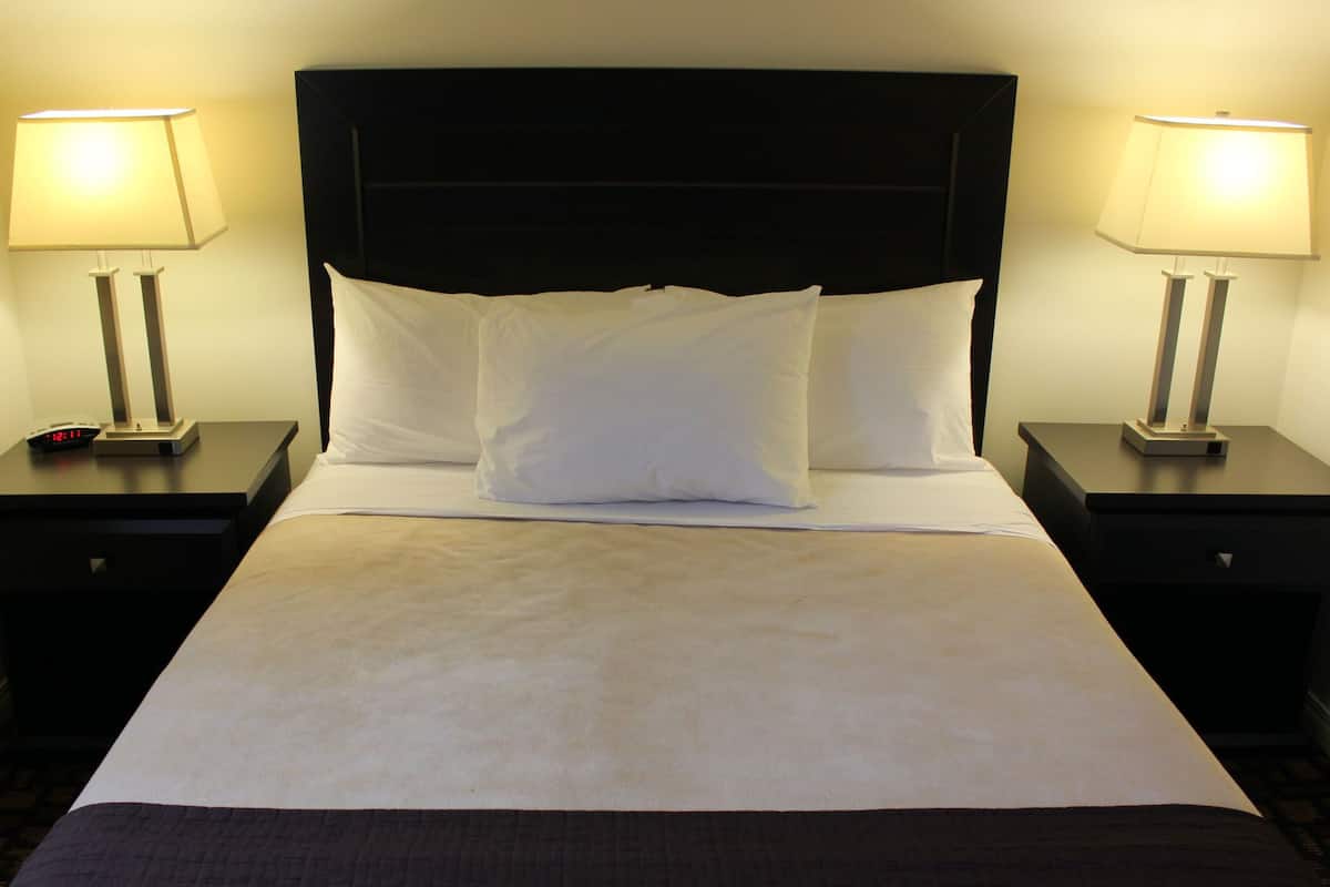 Uma cama bem arrumada com lençóis brancos e cabeceira escura. Há duas mesas de cabeceira, cada uma com uma luminária e um relógio.