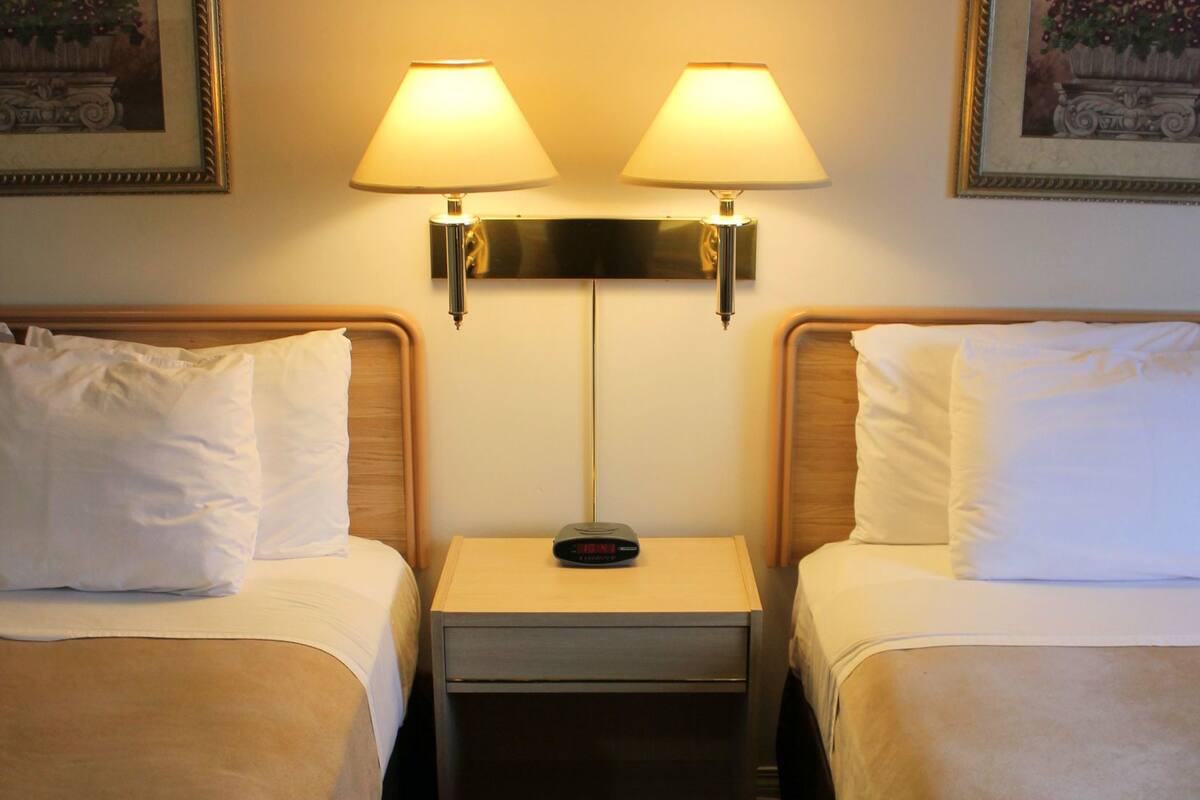 Quarto de hotel com duas camas, uma mesinha de cabeceira entre elas e duas luminárias na parede.