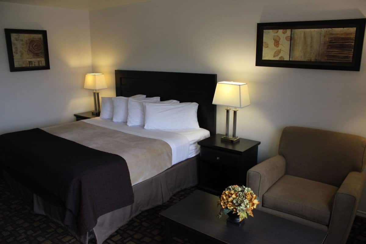 Quarto de hotel com uma cama, duas mesas de cabeceira com luminárias, uma cadeira, uma mesinha com um vaso e duas obras de arte emolduradas nas paredes.