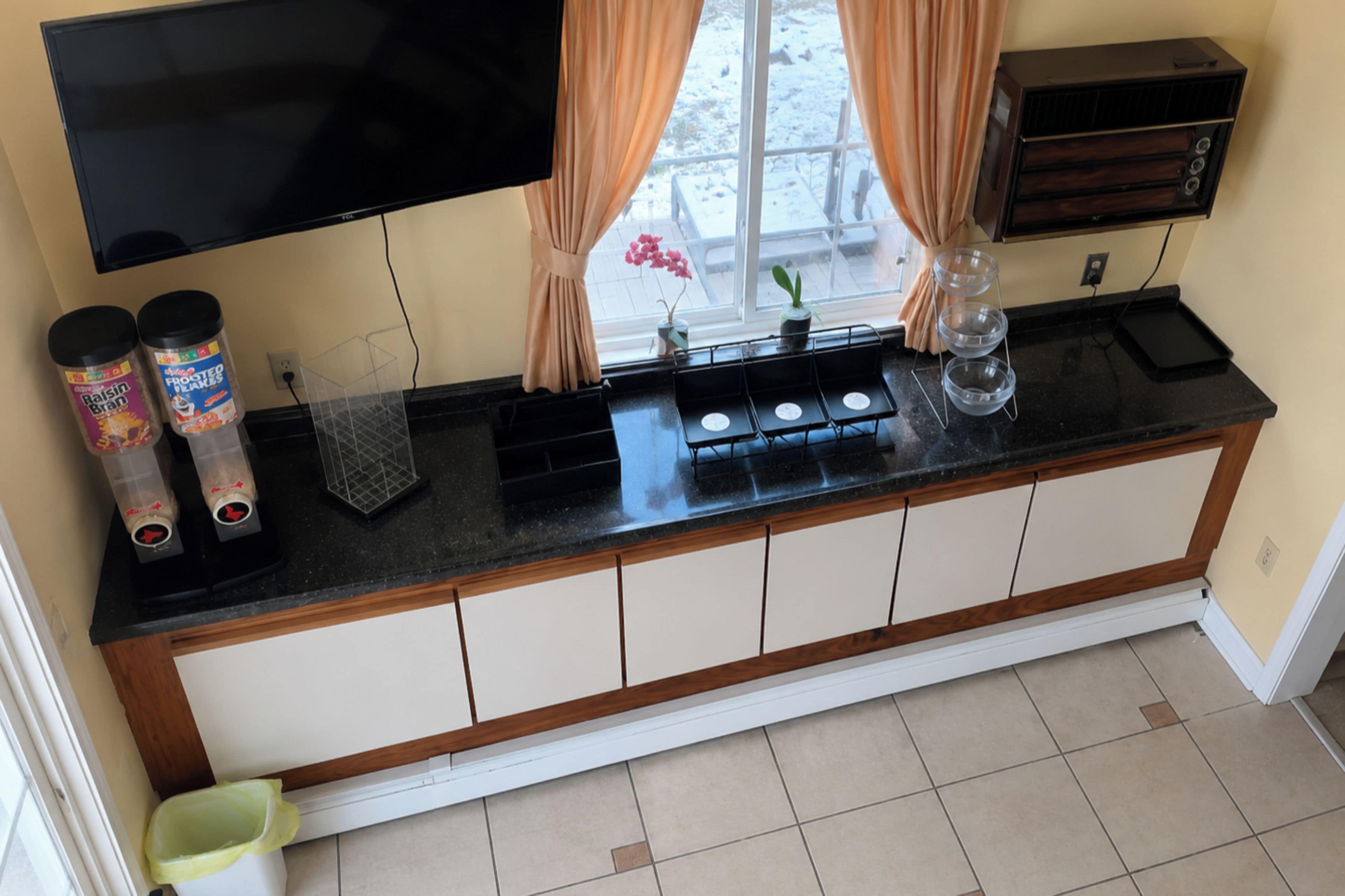 A kitchen with a black countertop, white cabinets, a TV mounted above, and a window with curtains.