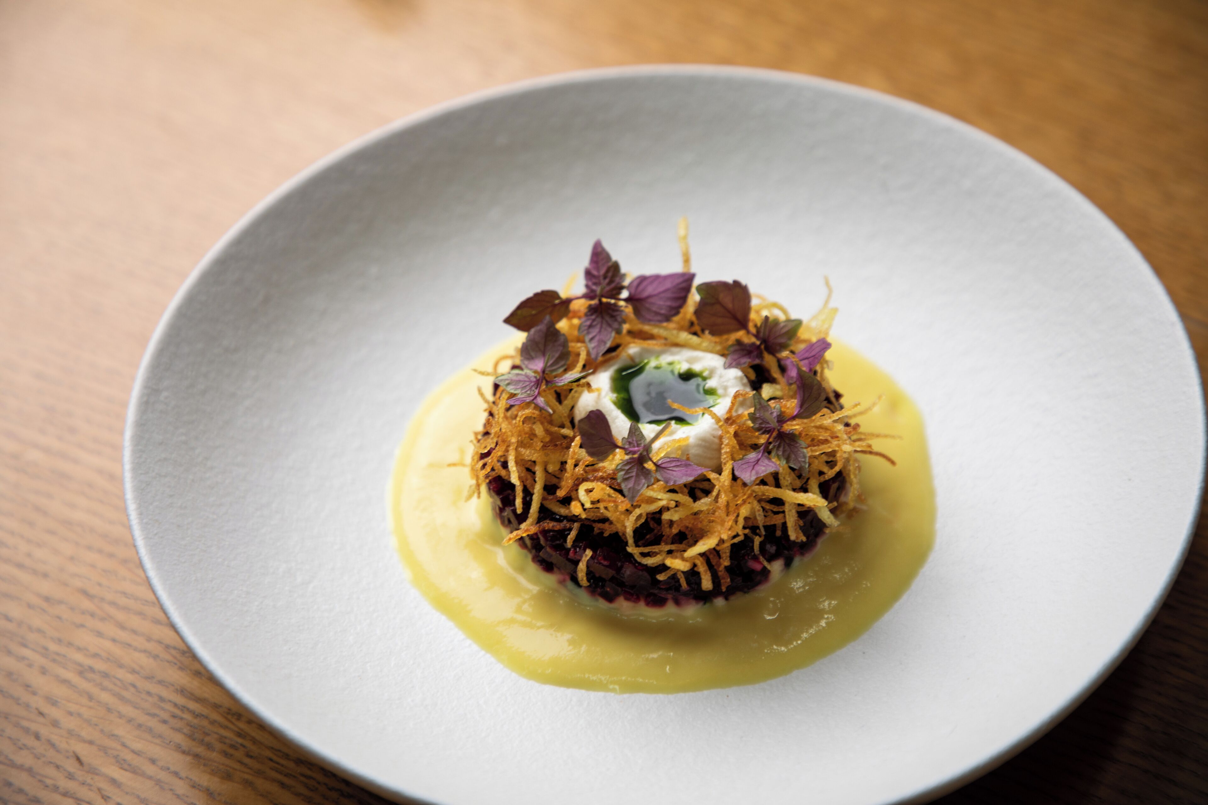 A white plate with a central food item, garnished with purple leaves and golden flakes, on a wooden table.