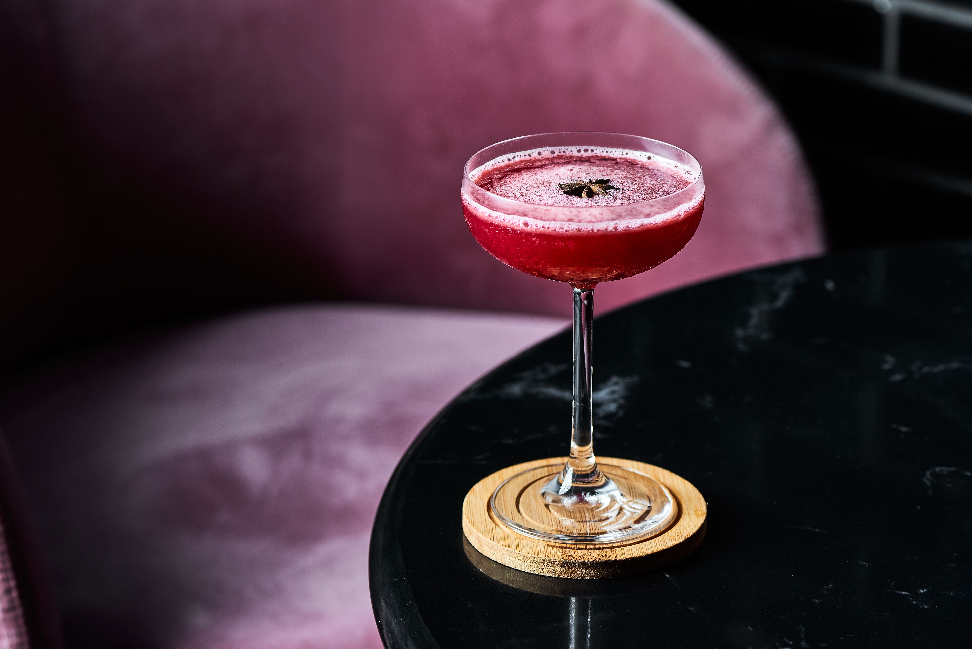 Un cóctel en un vaso sobre un posavasos de madera, sobre una mesa negra con un fondo rosa borroso.