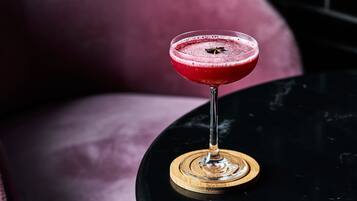 Un cóctel en un vaso sobre un posavasos de madera, sobre una mesa negra con un fondo rosa borroso.