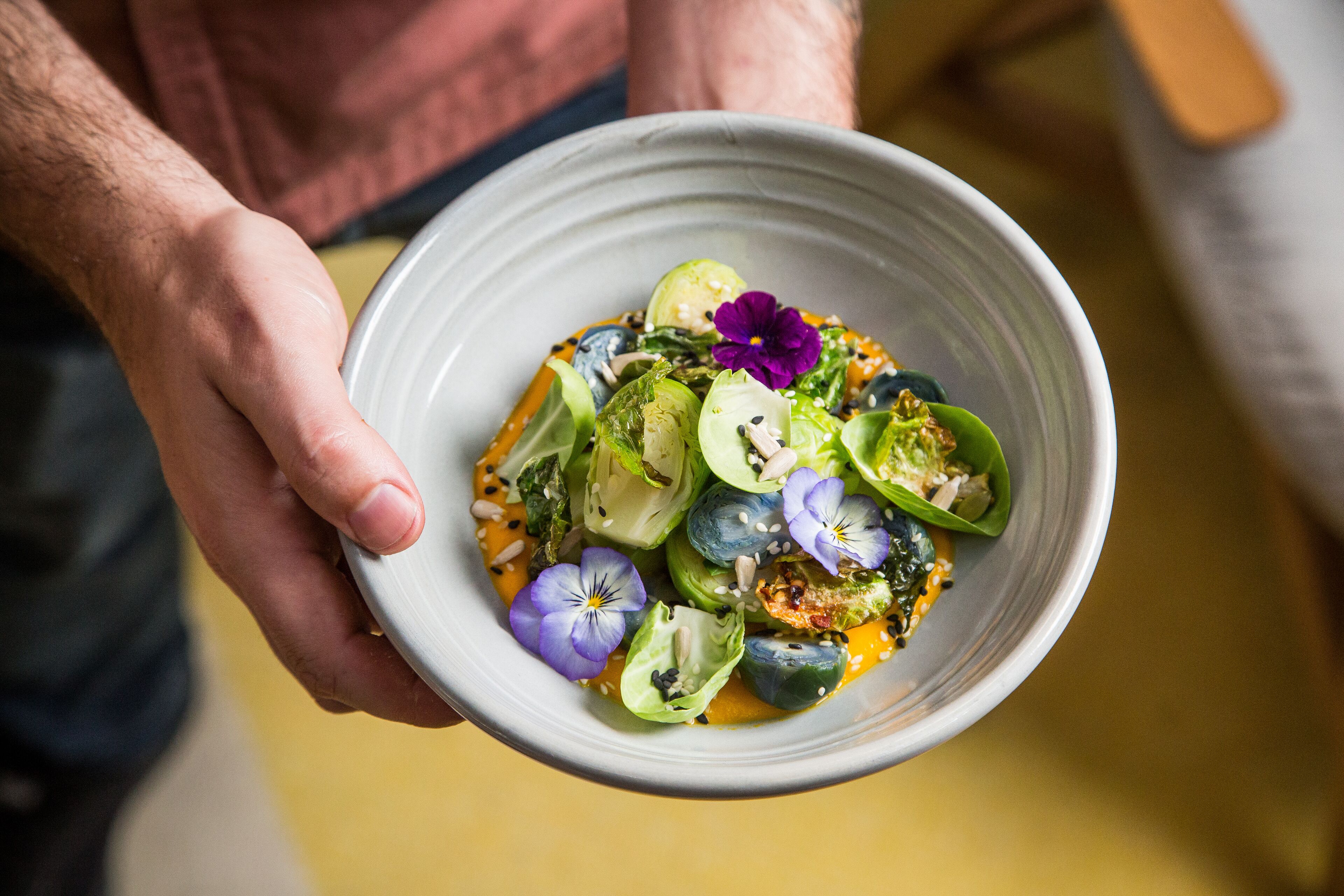 A person holding a bowl of food with various vegetables and flowers.