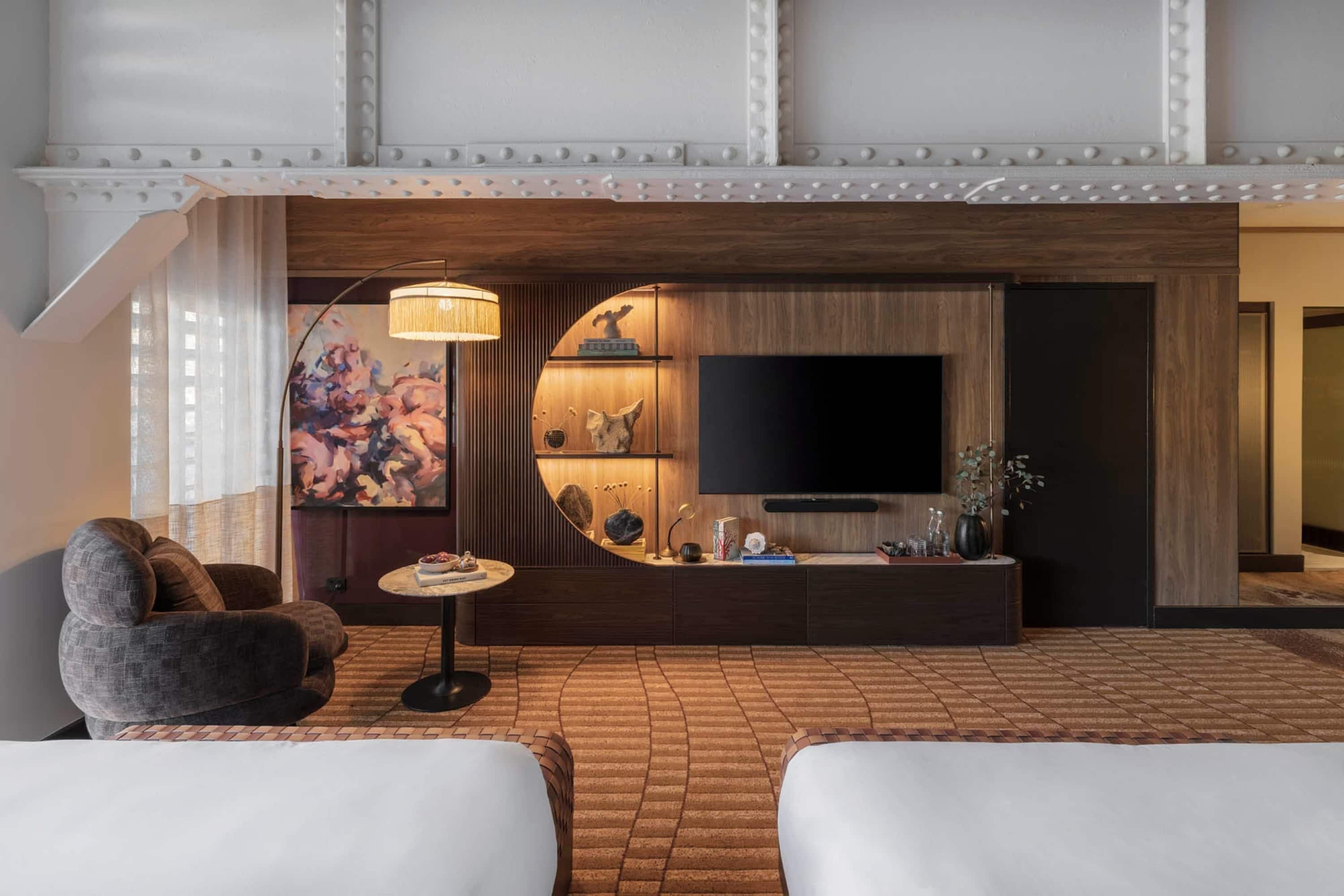A modern hotel room with a large flat-screen TV, a round wooden console table, a sofa, and a patterned floor.