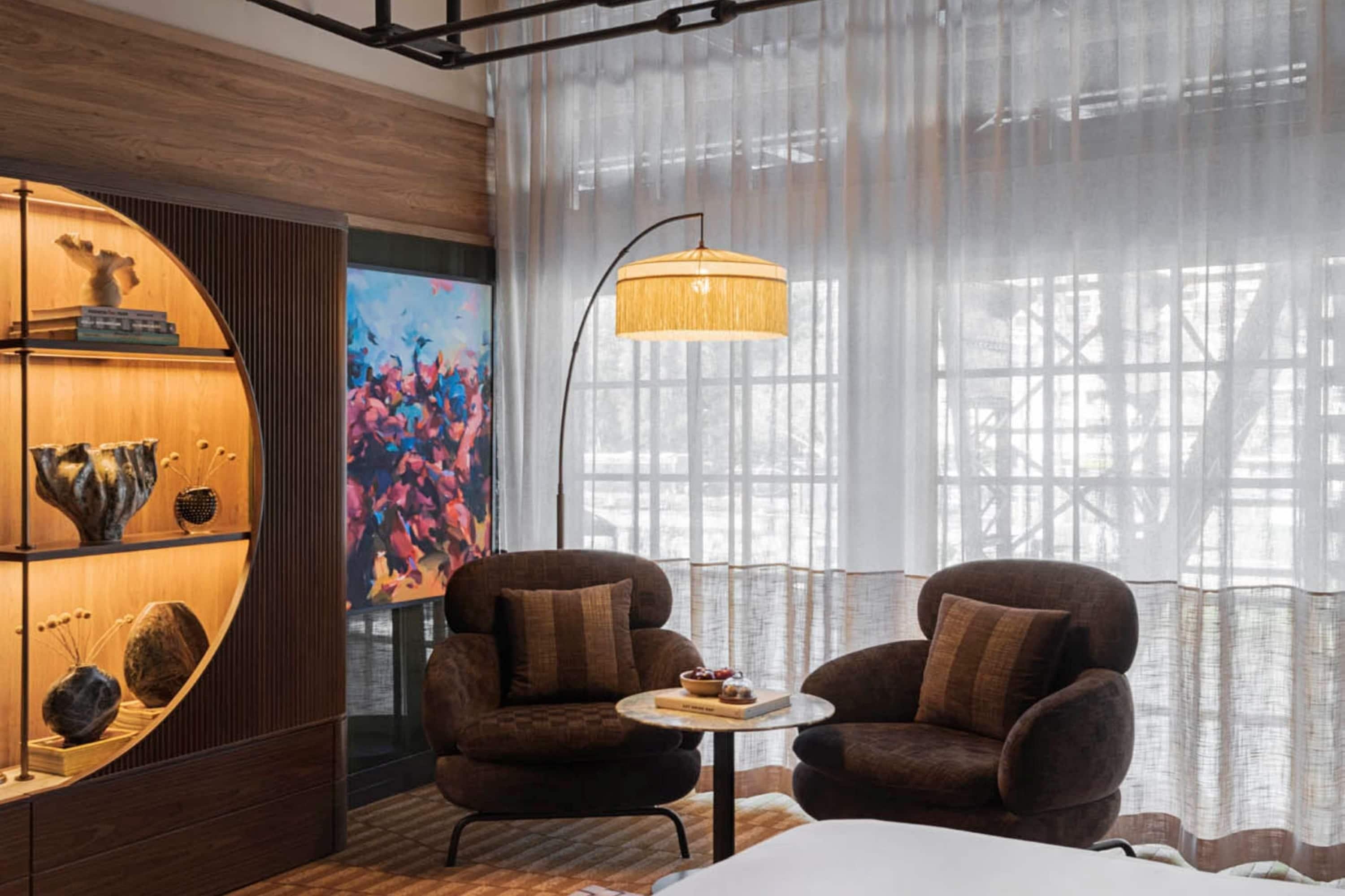 Una habitación moderna con dos sillones, una mesita redonda y un ventanal con cortinas translúcidas.