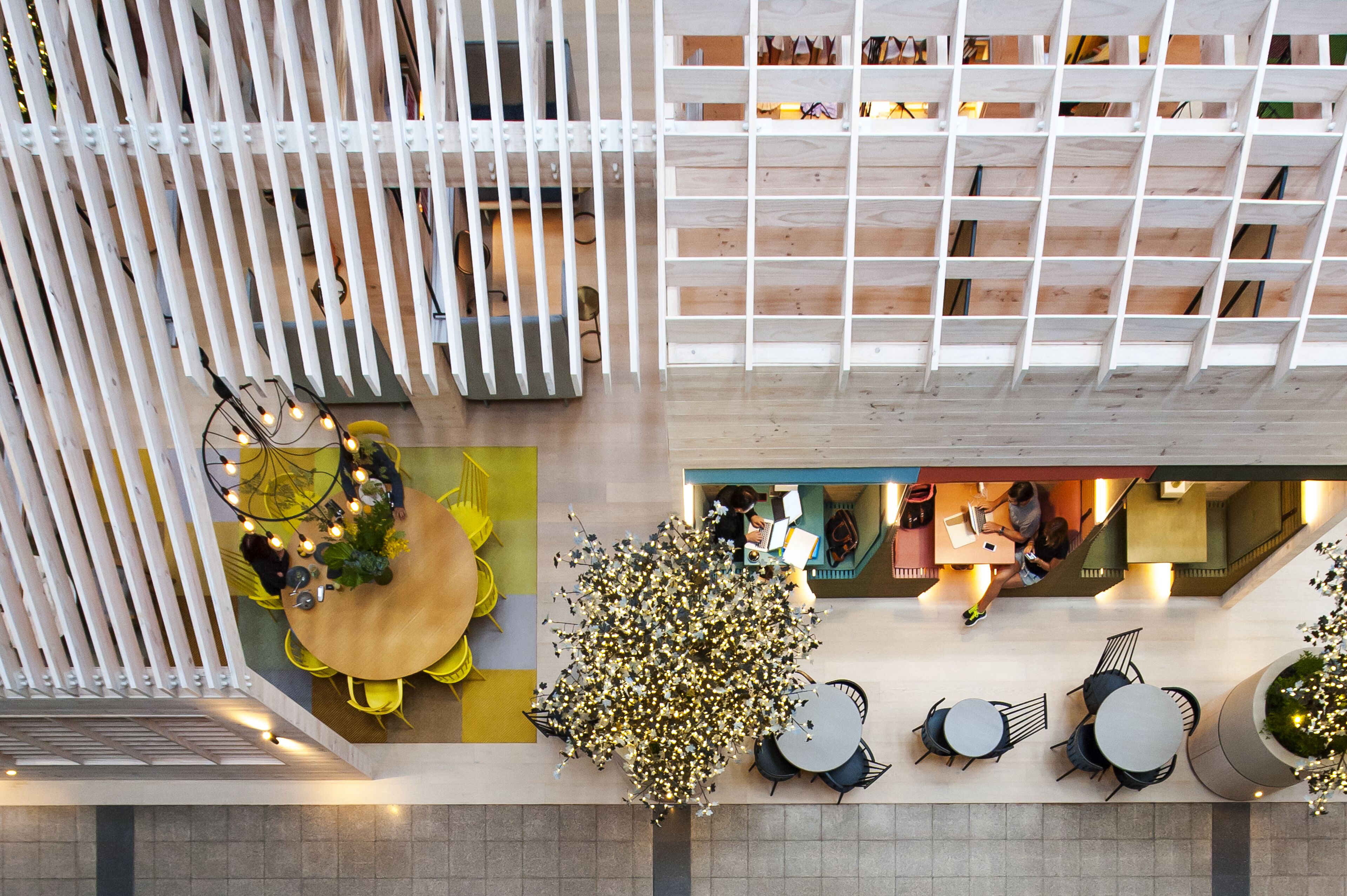 A modern indoor space with a wooden floor, a central round table with chairs, a seating area with yellow chairs, a white partition with shelves, and a staircase.