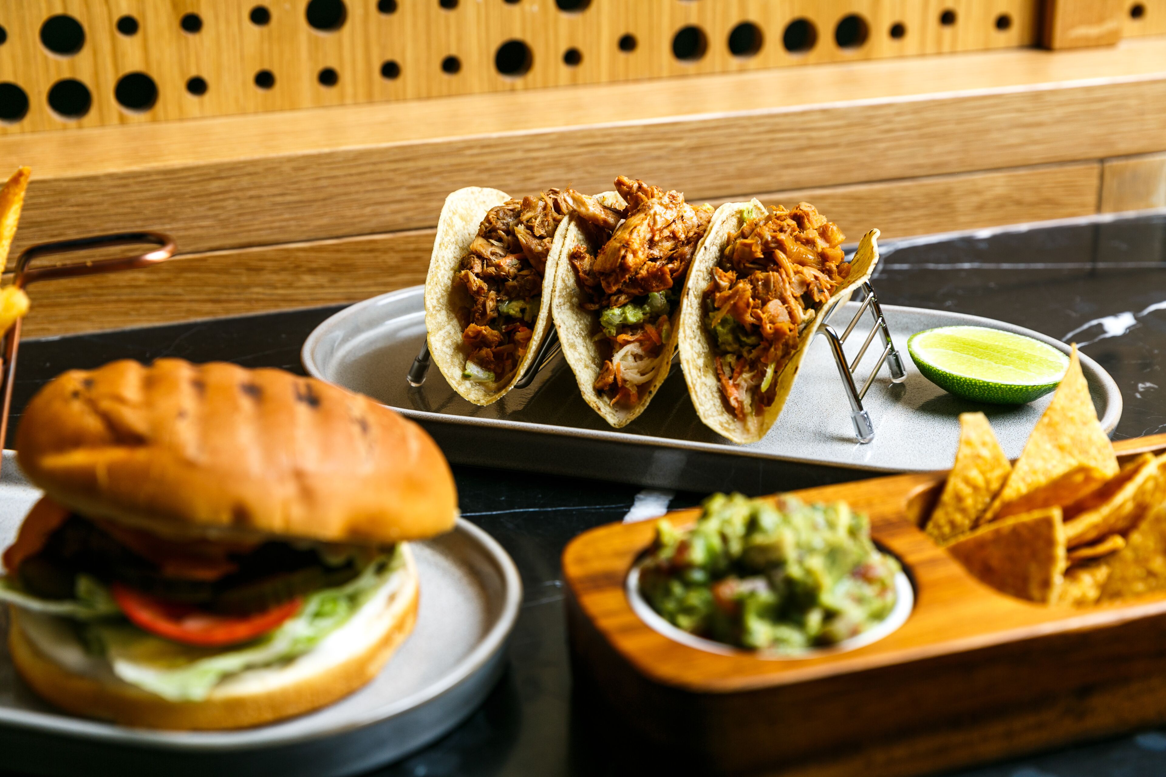 Una bandeja con tres tacos, una cuña de limón, un tazón de guacamole, un sándwich y totopos.