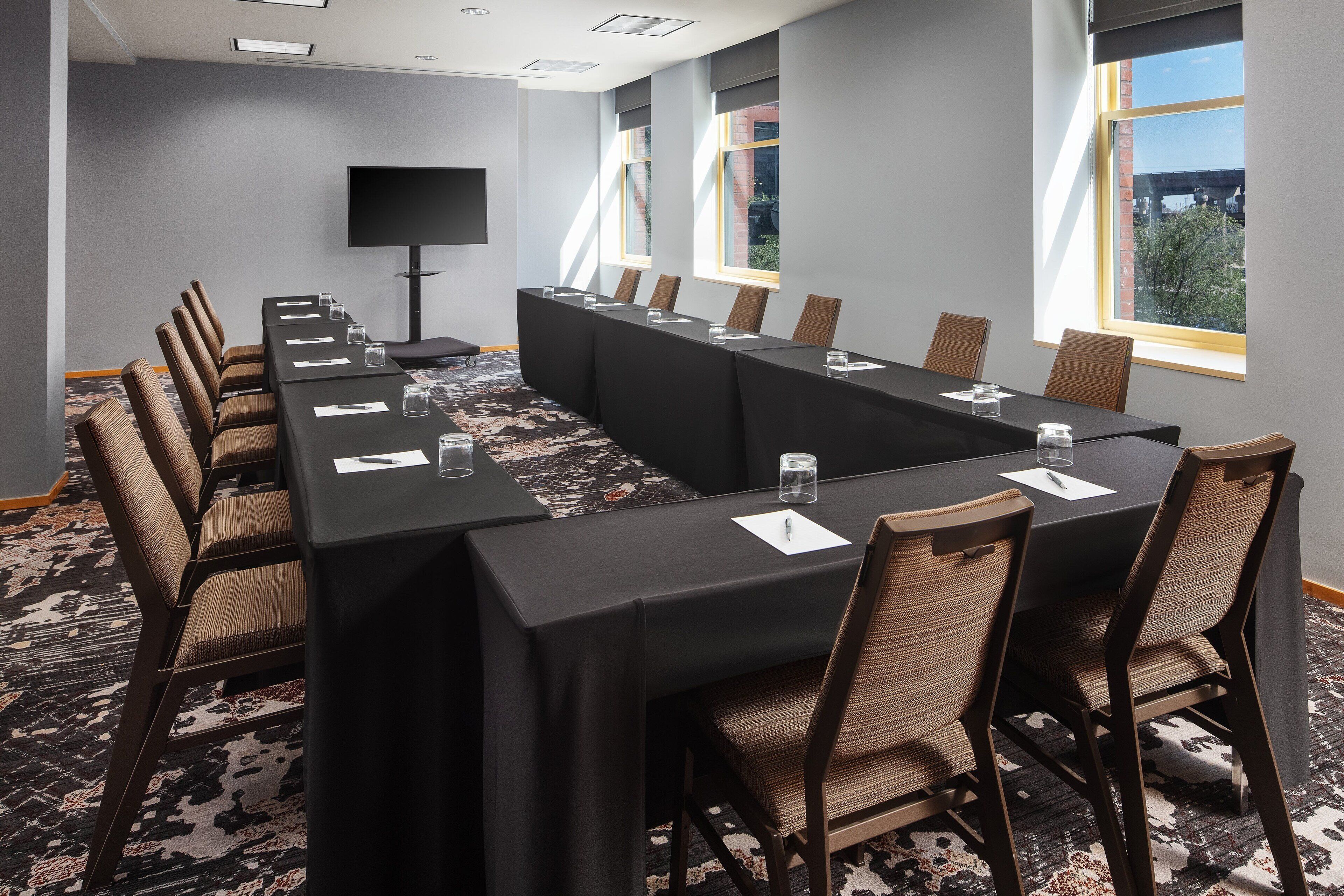 A conference room with a long table, chairs, a flat-screen TV, and large windows with a view of buildings and trees.