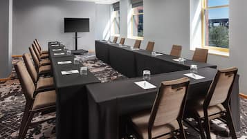 A conference room with a long table, chairs, a flat-screen TV, and large windows with a view of buildings and trees.