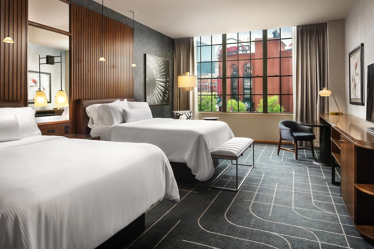 Ein Hotelzimmer mit zwei Betten, einem Schreibtisch, einem Stuhl, einem großen Fenster mit Blick auf ein Gebäude und einem gemusterten Teppich.
