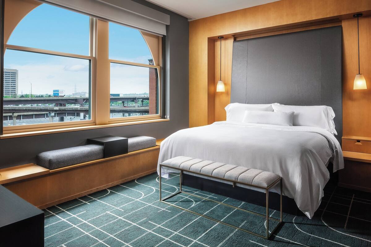 Ein modernes Hotelzimmer mit großem Fenster, einem Bett mit weißer Bettwäsche, einer Bank mit Kissen und Blick auf die Skyline der Stadt.