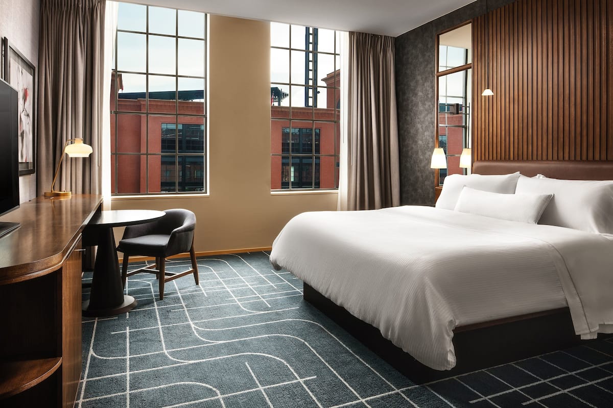 Ein Hotelzimmer mit einem großen Bett, einem Schreibtisch, einem Sessel und Blick auf eine Stadtlandschaft durch große Fenster.