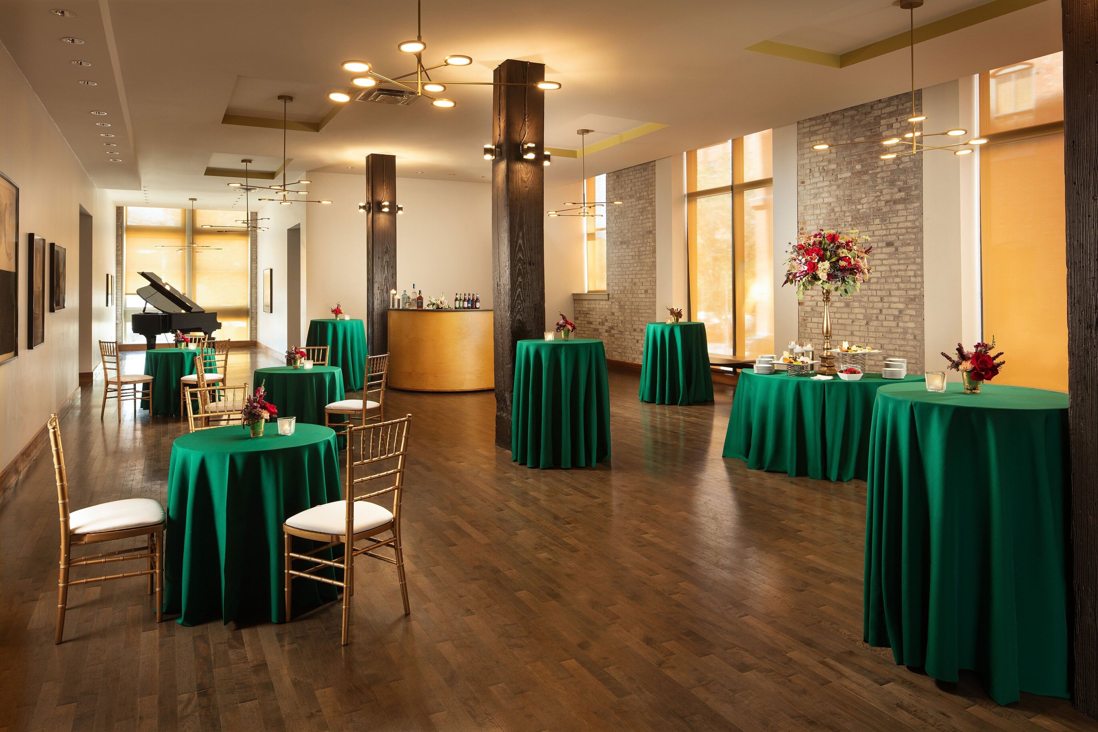 A spacious hall with green-covered tables and chairs, a grand piano, and a bar area.