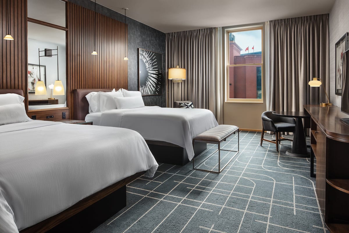 Ein Hotelzimmer mit zwei Betten, einem Schreibtisch, einem Stuhl, einer Lampe, einem Spiegel und Blick auf ein Gebäude.