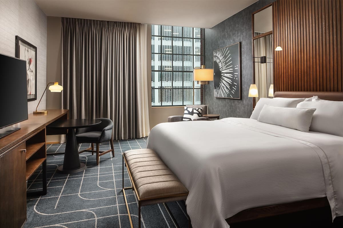 Ein Hotelzimmer mit einem großen Bett, einem Schreibtisch mit Stuhl, einer Bank, einem Fernseher und einem Fenster mit Vorhängen.