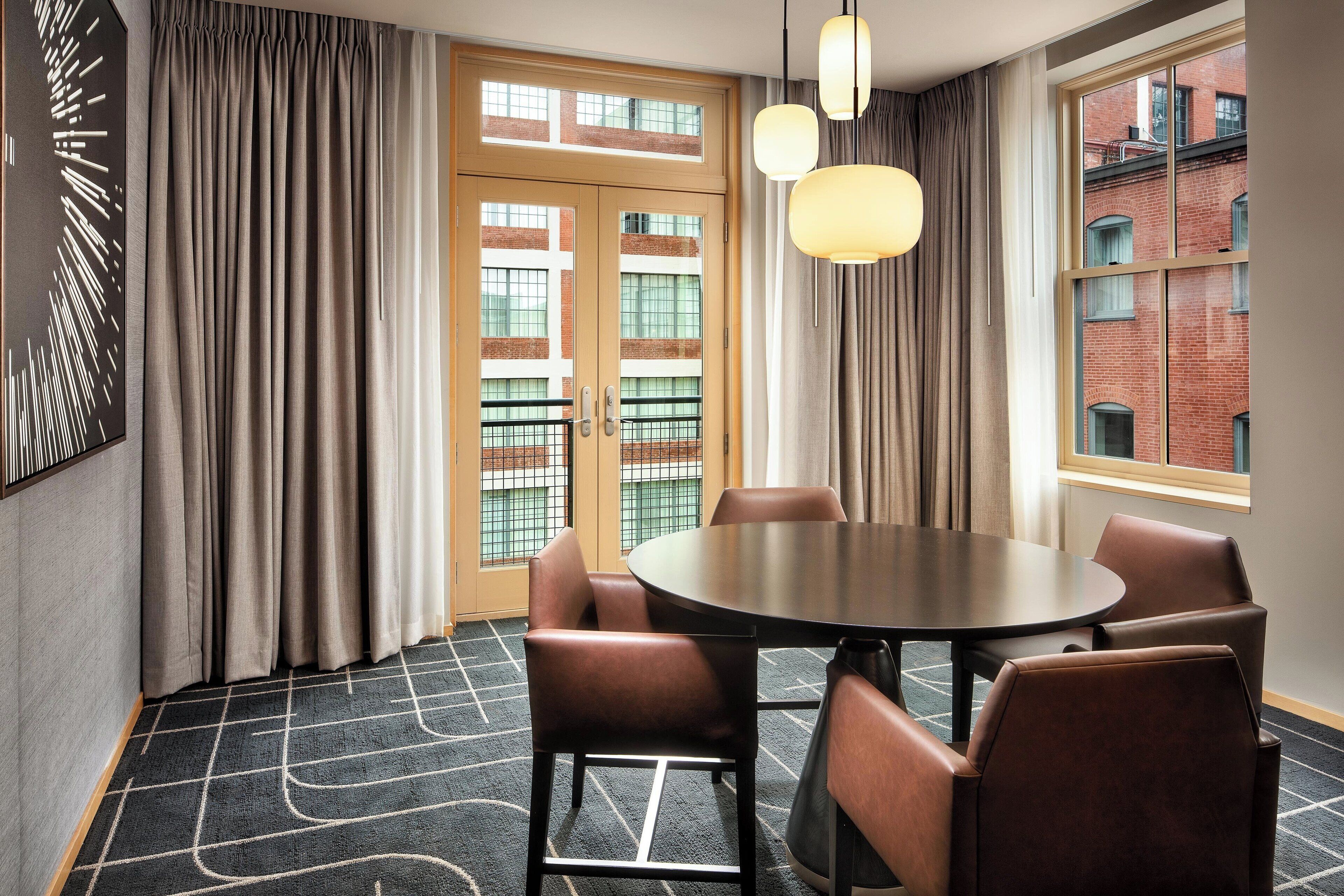 A conference room with a round table, six leather chairs, and a view of a brick building through large windows.