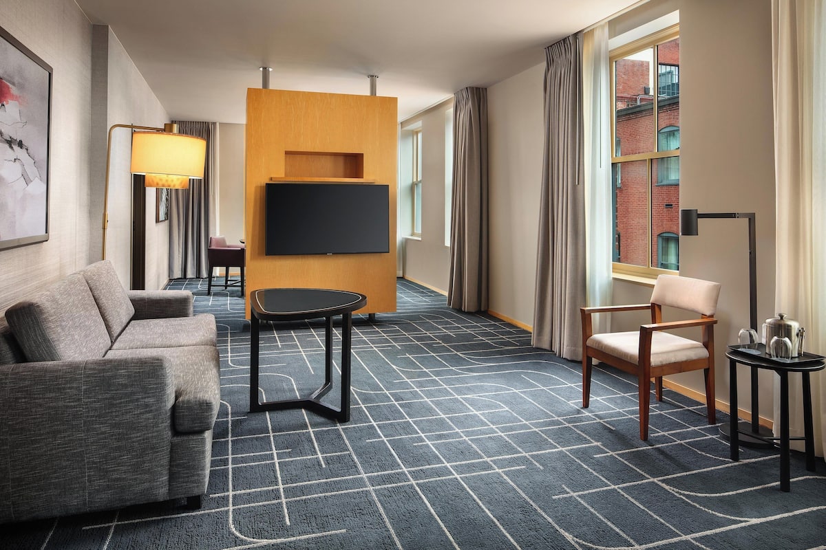 Ein Hotelzimmer mit einer Sitzecke, einem kleinen Tisch, einem an einer Holzvertäfelung befestigten Fernseher, einem Sessel und einem Fenster mit Vorhängen.