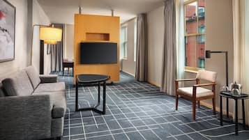 A hotel room with a sofa, a small table, a TV mounted on a wooden panel, a chair, and a window with curtains.