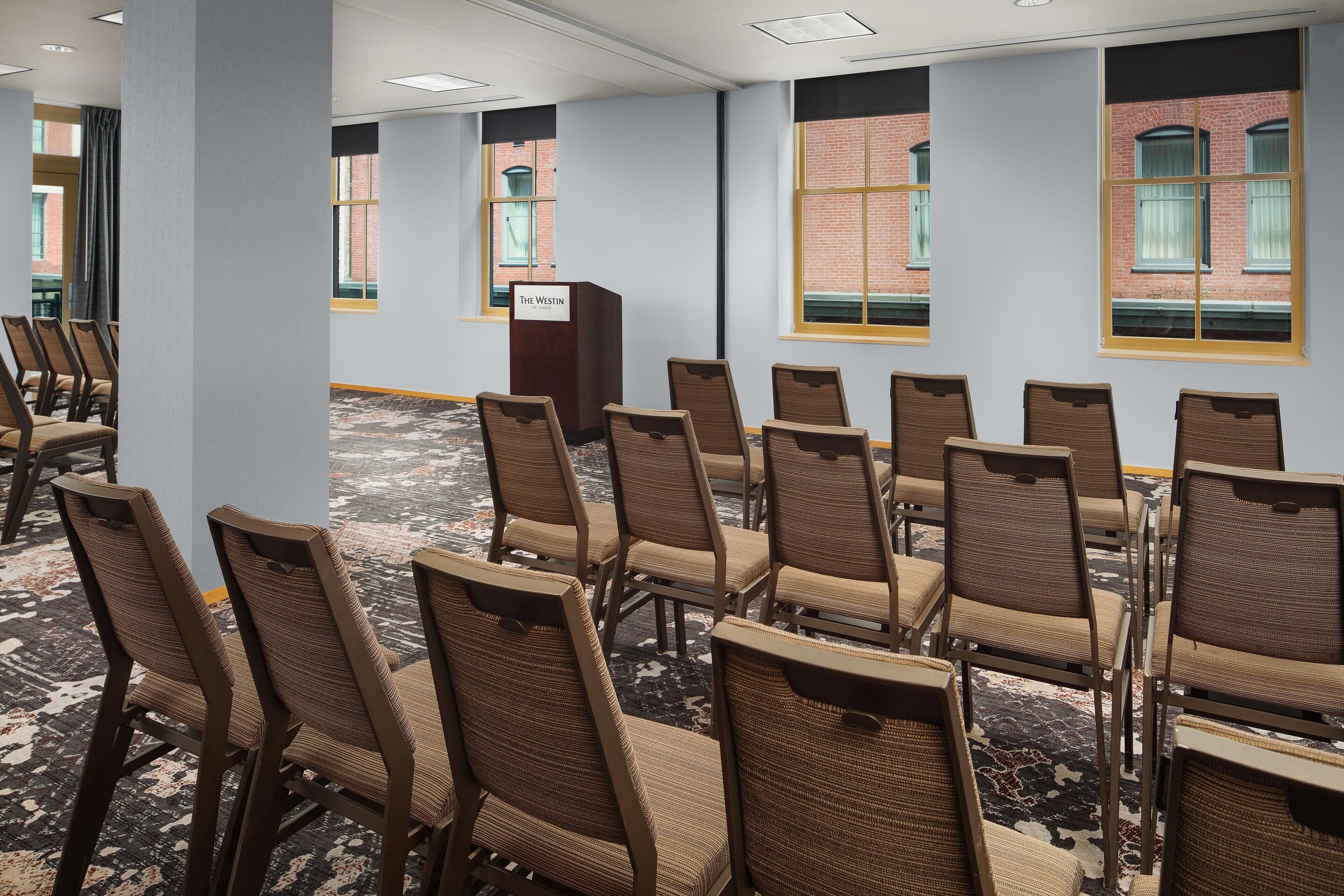 A conference room with rows of chairs, a podium, and large windows with a brick wall view.