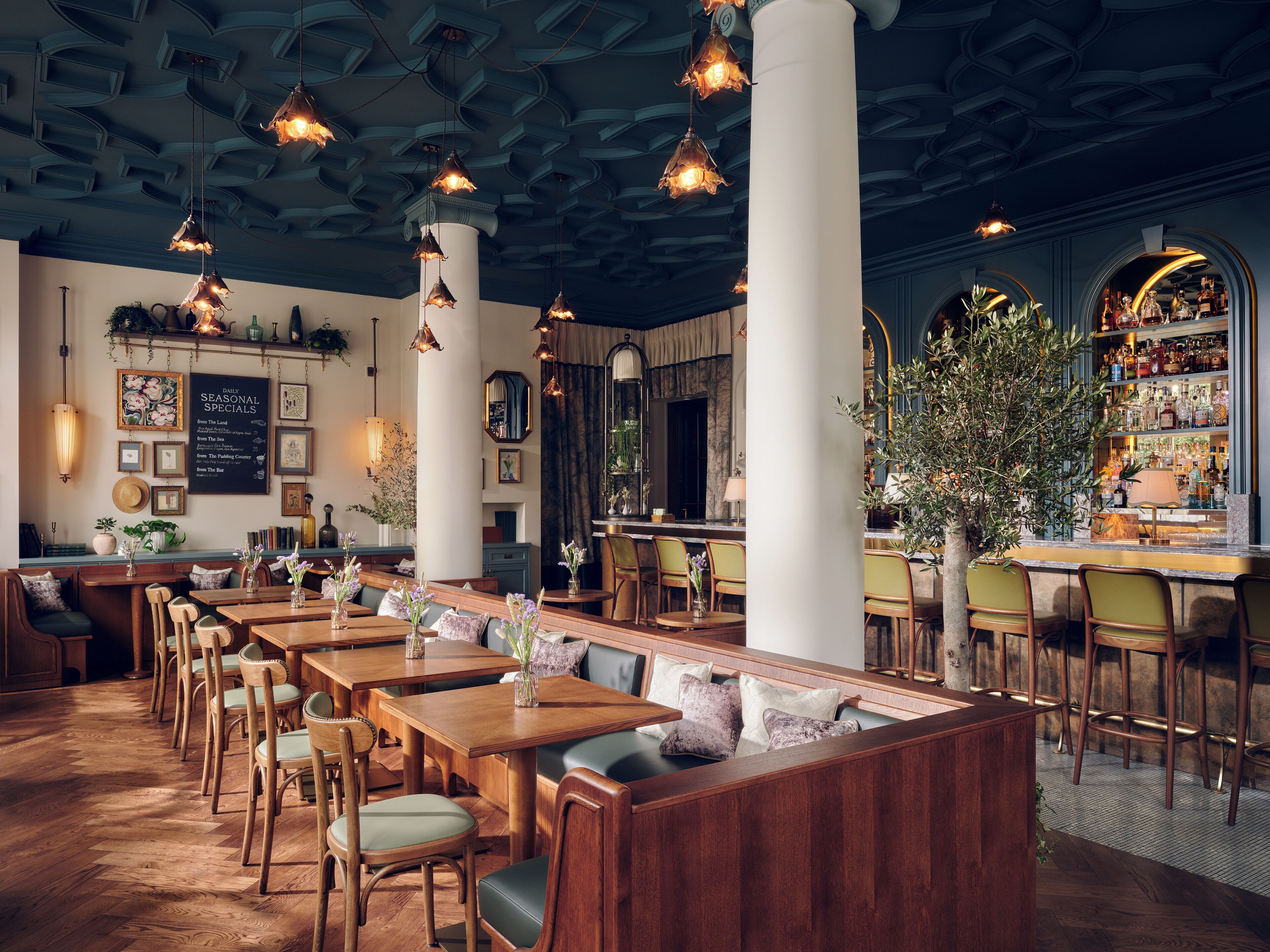 A restaurant interior with wooden tables, chairs, hanging pendant lights, a bar area, and a menu board.