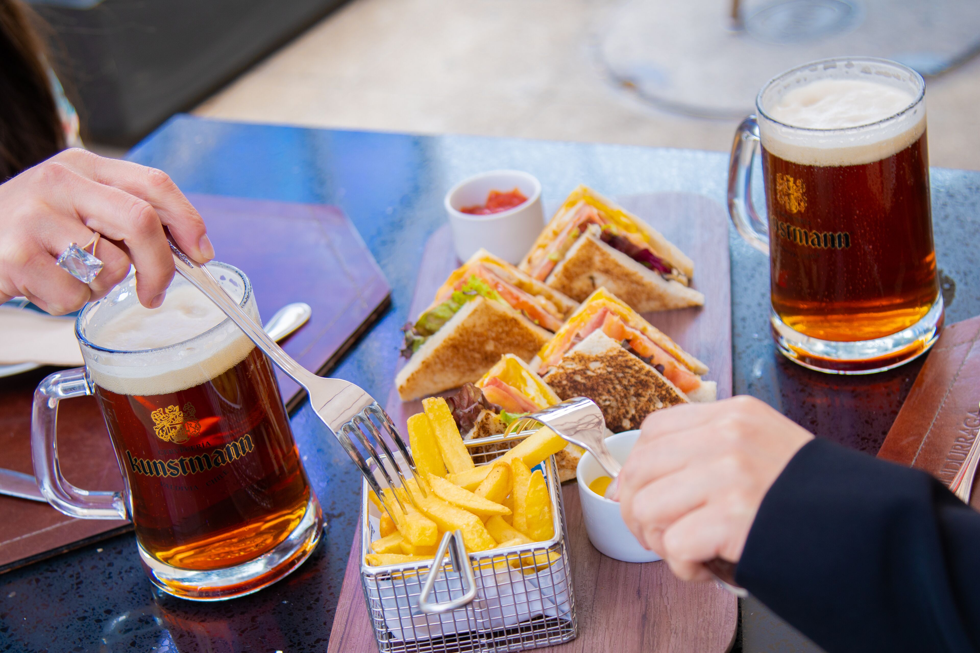Uma mesa com duas canecas de cerveja, um cesto de batatas fritas e sanduíches.