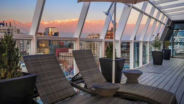A rooftop terrace with wicker lounge chairs, potted plants, and a view of a cityscape and mountains.