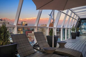 Terraza en la azotea con sillones de mimbre, plantas en macetas y vistas a un paisaje urbano y montañas.