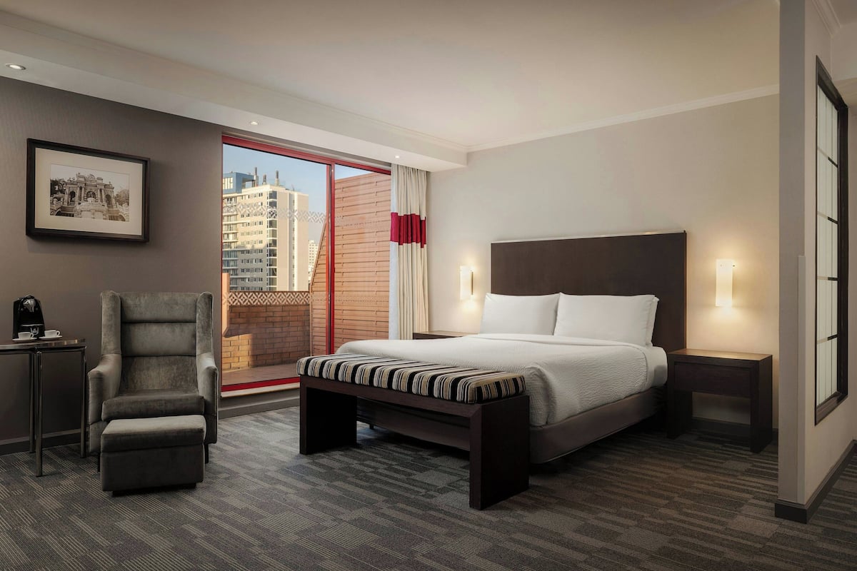 Hotelkamer met een bed, een stoel, een bankje, een nachtkastje, een aan de muur hangend schilderij en uitzicht op gebouwen door een groot raam.