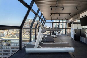 Un gimnasio con cintas de correr y máquinas de pesas, ventanas amplias y vistas a la ciudad.